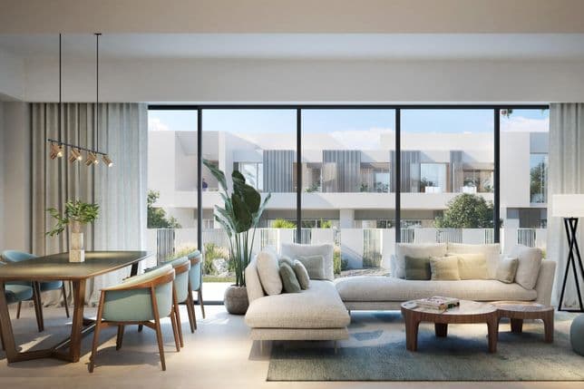 Modern living room with large windows, beige sectional sofa, wooden tables, and dining area. Outside view of contemporary buildings.