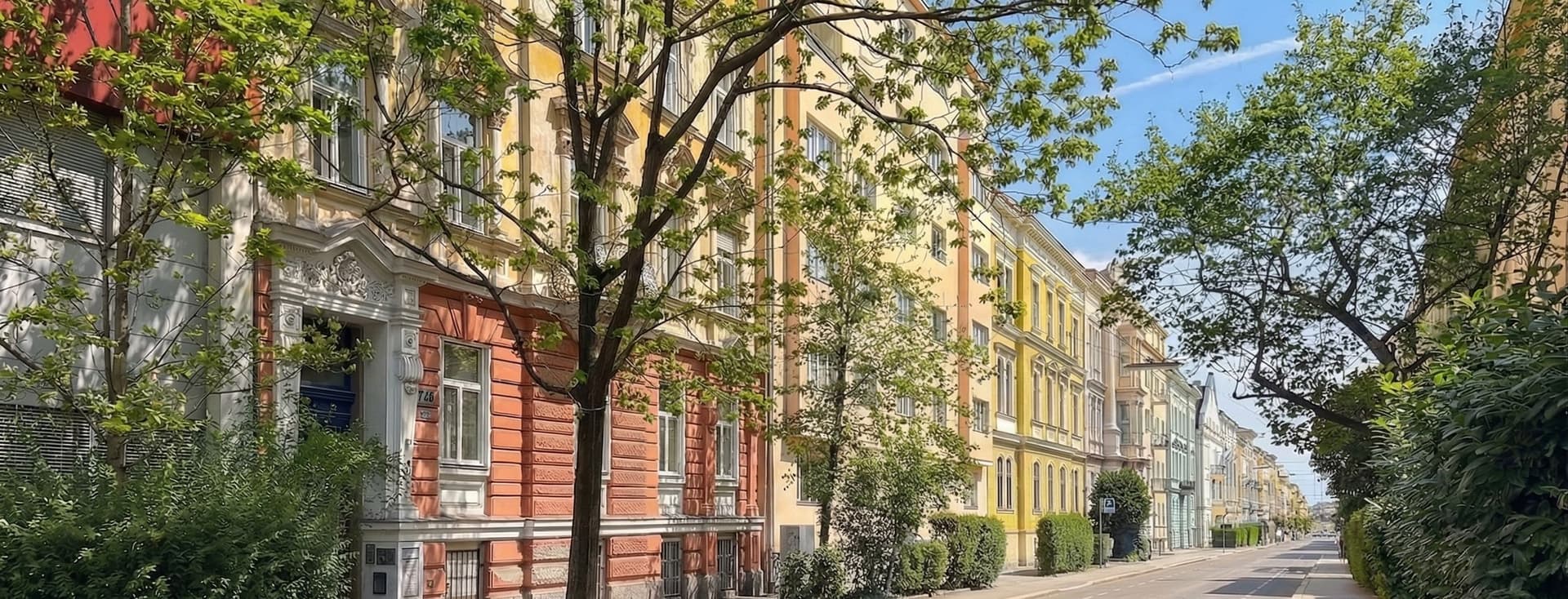 Straßenansicht mit farbenfrohen Gebäuden im europäischen Stil und Bäumen, die den Bürgersteig säumen, an einem sonnigen Tag.