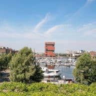 View of Antwerp harbor with boats, green trees, and the MAS museum, under a blue sky.
