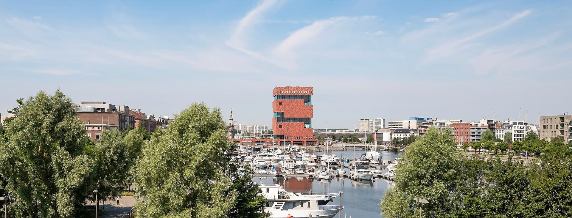 View of Antwerp harbor with boats, green trees, and the MAS museum, under a blue sky.