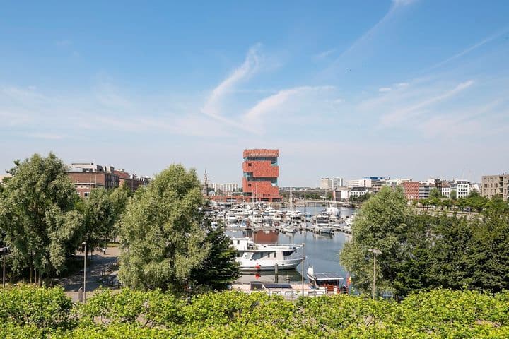 Vue sur le port d'Anvers avec des bateaux, des arbres verts et le musée MAS, sous un ciel bleu.