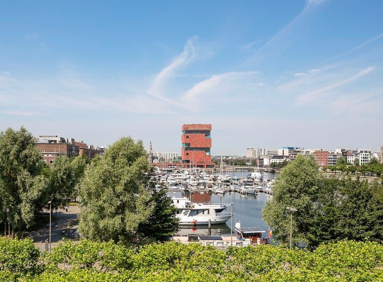 Uitzicht op de haven van Antwerpen met boten, groene bomen en het MAS museum, onder een blauwe lucht.