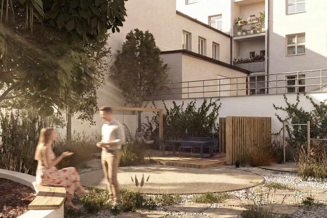 Landscaped residential courtyard with abundant greenery, ornamental grasses, and a mature tree. In the foreground, an outdoor table with metal chairs stands on a wooden deck. A pathway leads to a building entrance with a small balcony above the door. On the right, a woman sits on a bench beneath the tree, while another person walks down the entrance steps. The atmosphere feels calm and community-oriented.