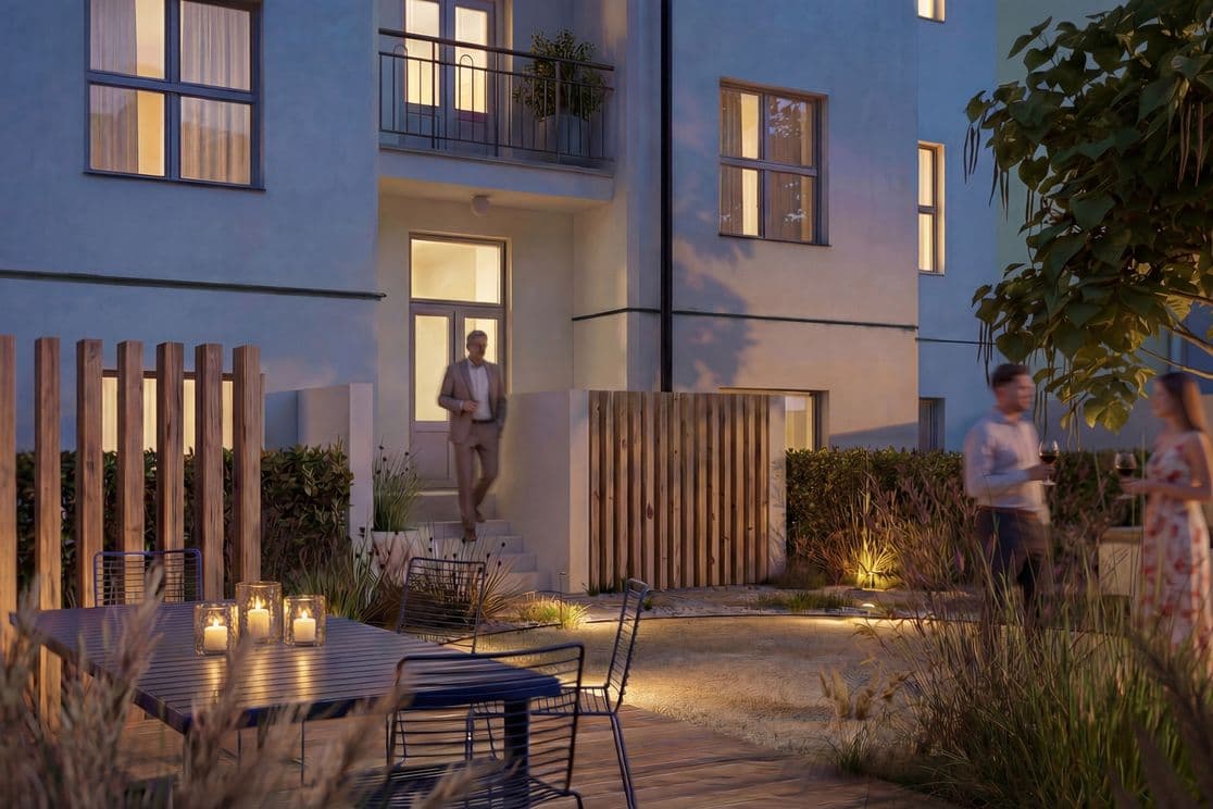 Evening view of a residential courtyard with landscaped greenery, ornamental grasses, and a mature tree. In the foreground, an outdoor table with metal chairs is decorated with glowing candle lanterns. Illuminated pathways lead to the building entrance, where a man is walking down the steps. On the right, two people stand holding glasses of wine. Lit apartment windows and outdoor lighting create a warm and inviting atmosphere.
