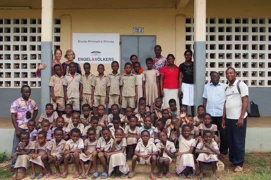 A group of children and adults pose in front of a school building with a sign reading "École Primaire Privée." Everyone is smiling.