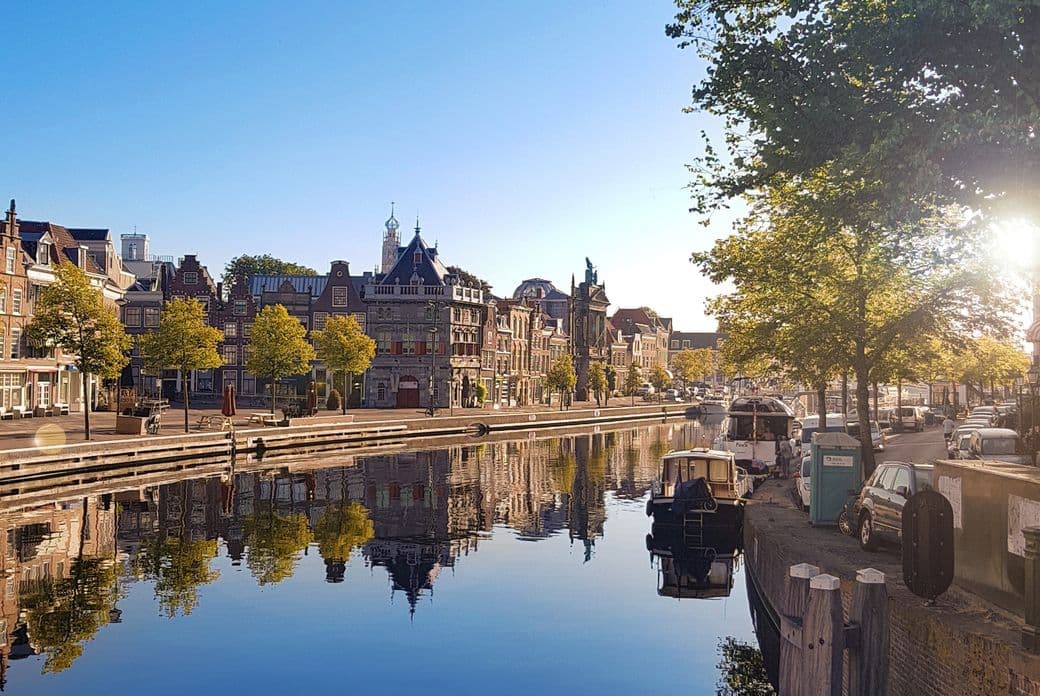 Waterfront view of Haarlem, Netherlands, with historic buildings reflected in the canal under a clear blue sky. Boats are docked along the quay.