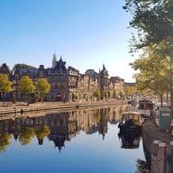 Waterfront view of Haarlem, Netherlands, with historic buildings reflected in the canal under a clear blue sky. Boats are docked along the quay.