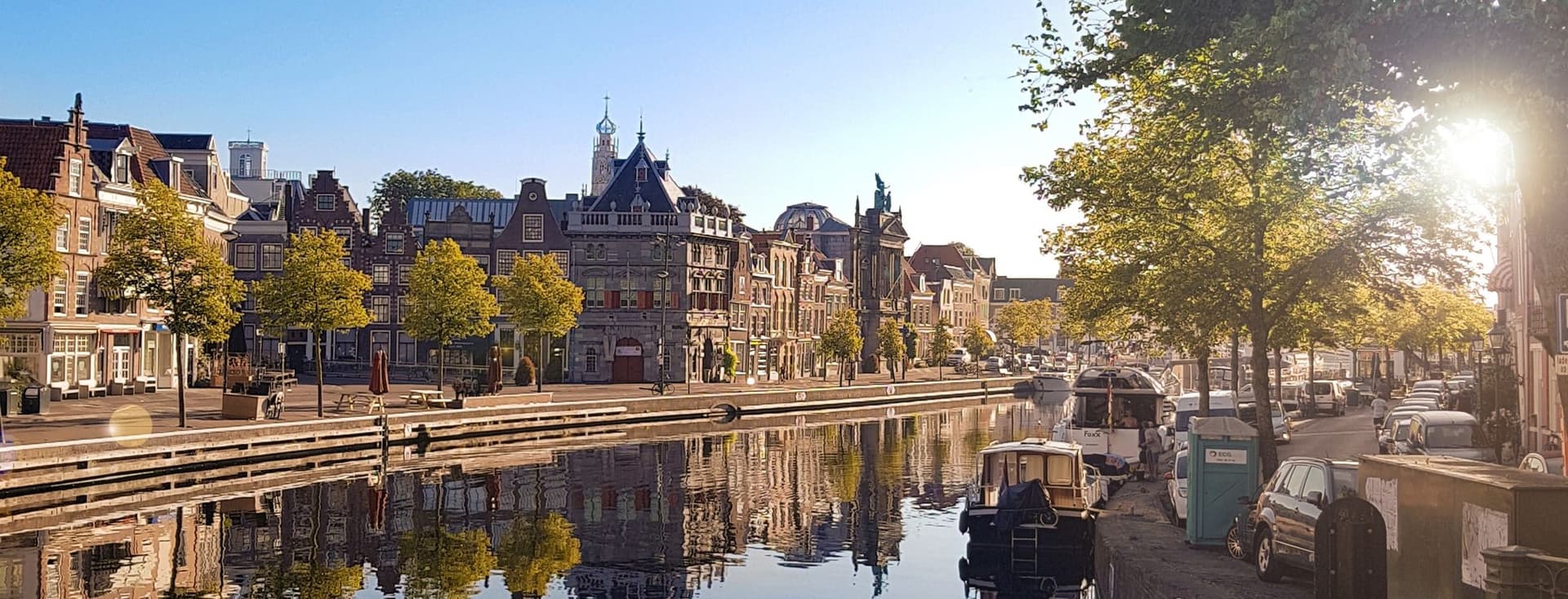 Waterfront view of Haarlem, Netherlands, with historic buildings reflected in the canal under a clear blue sky. Boats are docked along the quay.