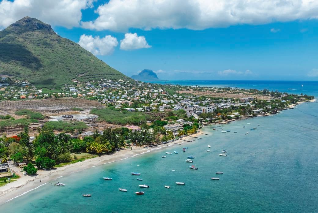 Aerial view of Tamarin with boats on turquoise water, lush greenery, and a mountain in the background under a partly cloudy sky.