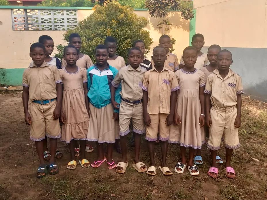 Group of students in beige uniforms standing outside a school, wearing sandals, with a wall and greenery in the background.