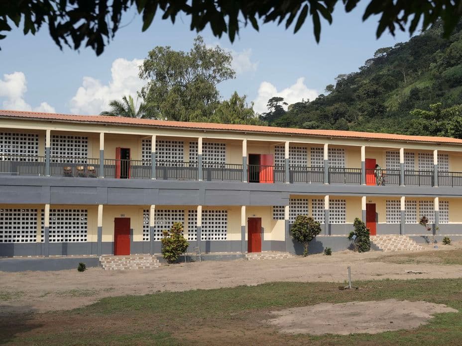 A two-story school building with red doors and grid-patterned windows, surrounded by trees and a grassy field, under a partly cloudy sky. The school was build by Engel & Völkers Charity