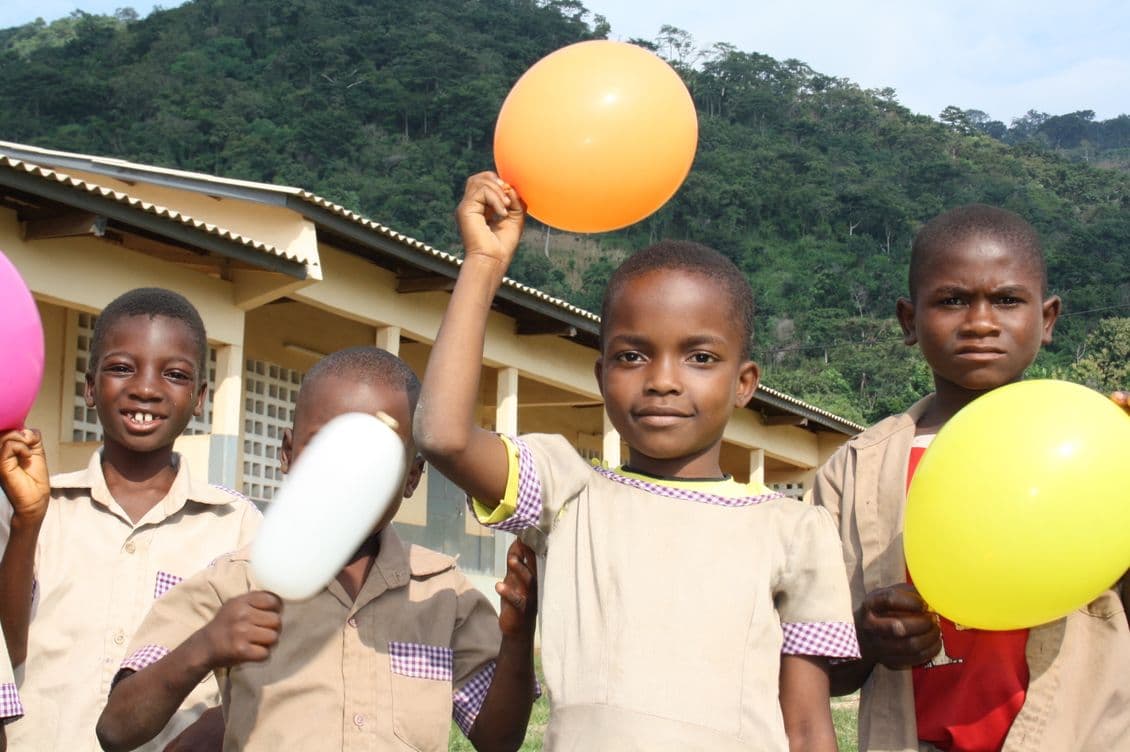 Children in school uniforms holding balloons, smiling in front of a building with green hills in the background.