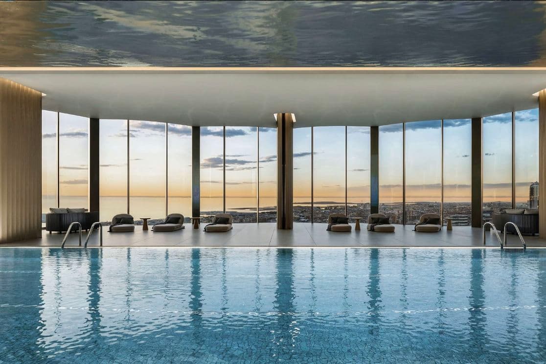 Indoor infinity pool with a cityscape view through floor-to-ceiling windows, featuring lounge chairs under a reflective ceiling.