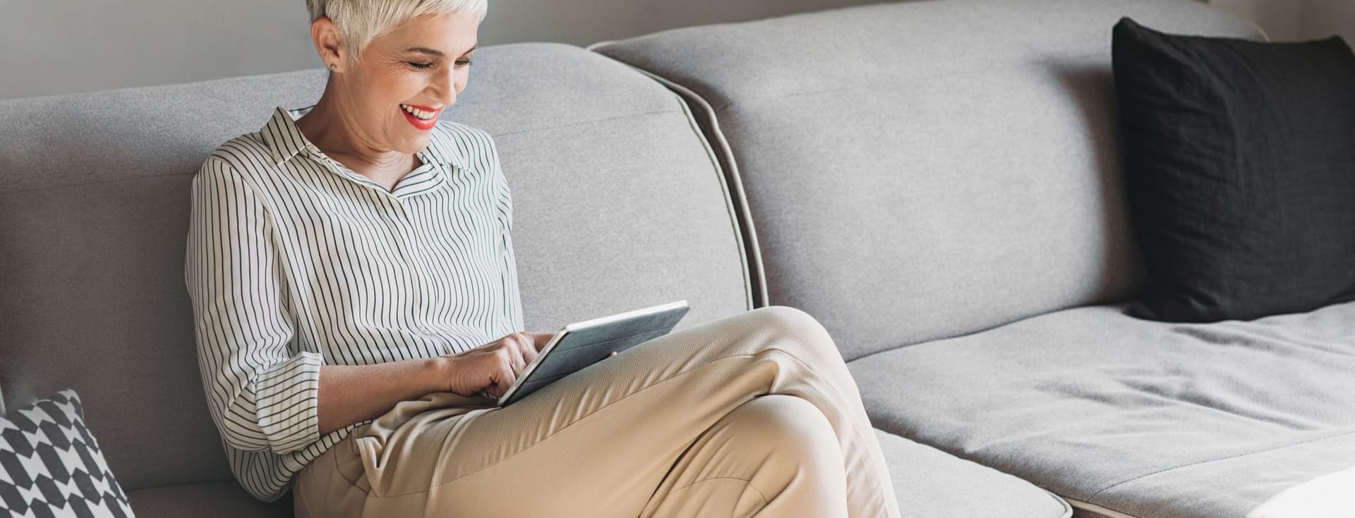 A smiling woman with short white hair sits on a gray sofa, using a tablet. She wears a striped shirt and khaki pants. A black pillow is on the sofa.