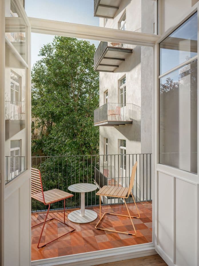 View from the interior onto a small balcony with terracotta tiles, furnished with two simple chairs and a round table. The balcony faces a green courtyard, with the building façade and neighboring balconies visible opposite.