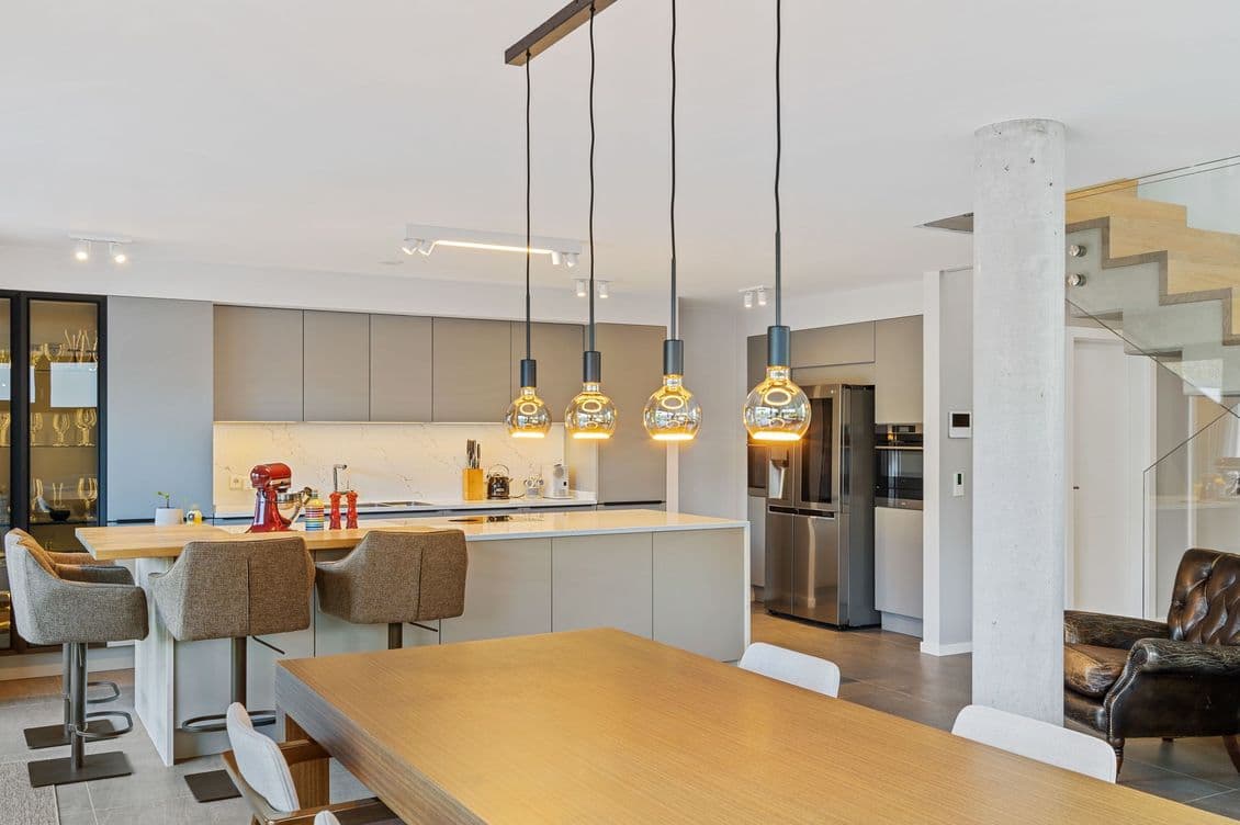 Modern kitchen and dining area with a wooden table, beige chairs, hanging pendant lights, and a sleek kitchen island with bar stools.