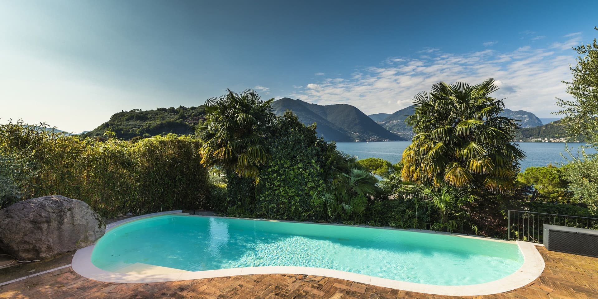 Ein ruhiger Pool, umgeben von üppigem Grün, bietet unter einem teilweise bewölkten Himmel Aussicht auf einen See mit Bergen in der Ferne.
