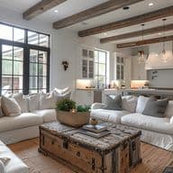 Ein helles, luftiges Wohnzimmer und eine Küche mit offenem Konzept im modernen Bauernhausstil. Der Raum verfügt über weiße Sofas mit Husse, die um einen rustikalen Holztruhen-Couchtisch angeordnet sind, freiliegende Holzbalken an der Decke, große schwarz gerahmte Fenster, einen natürlichen Juteteppich und eine angrenzende weiße Küche mit Pendelleuchten. Die neutrale Farbpalette umfasst Weiß, Creme und natürliche Holztöne mit Akzenten von Grün in einem Korb als Mittelpunkt.