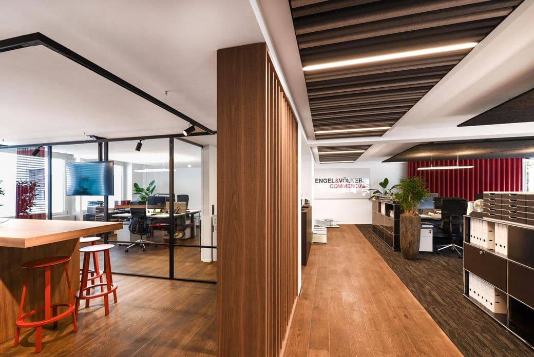 Modern office space with wooden flooring, a glass-walled conference room, desks, shelves, and red bar stools near a high table.