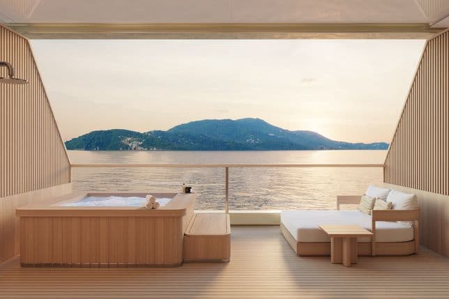 Ein luxuriöses Spa-Deck auf einer Holzyacht mit sprudelndem Whirlpool, Lounge-Sitzgelegenheiten und Handtüchern, von dem aus man bei Sonnenuntergang den Blick über das ruhige Meer auf eine grüne Insel genießen kann.