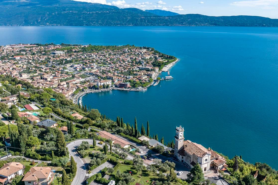 Aerial view of a lakeside town with a church, surrounded by lush greenery and mountains, overlooking a vast blue lake.