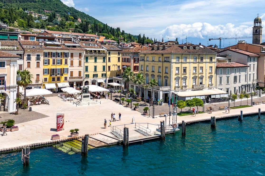 Vista panoramica di una città lacustre con edifici colorati, un vivace lungomare e montagne sullo sfondo sotto un cielo parzialmente nuvoloso.
