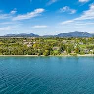 Luftaufnahme einer idyllischen Seelandschaft mit üppigem Grün, verstreuten Gebäuden und fernen Bergen unter einem blauen Himmel mit Wolken.