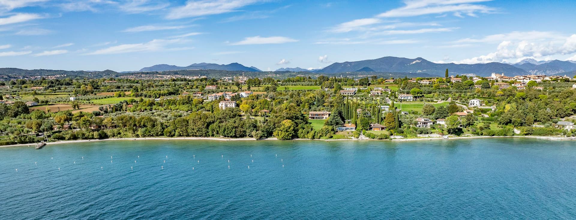 Luftaufnahme einer idyllischen Seelandschaft mit üppigem Grün, verstreuten Gebäuden und fernen Bergen unter einem blauen Himmel mit Wolken.