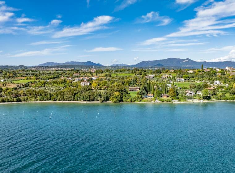 Luftaufnahme einer idyllischen Seelandschaft mit üppigem Grün, verstreuten Gebäuden und fernen Bergen unter einem blauen Himmel mit Wolken.