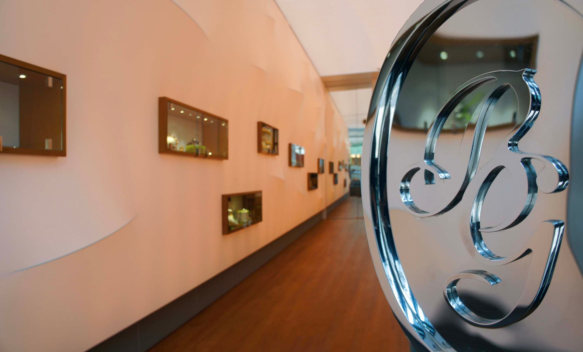Hallway view with wood floors, peach walls, and display cases. A large, shiny metal logo is in the foreground.