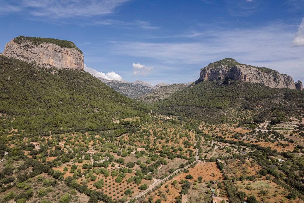 Vista aèria de dues grans muntanyes rocoses envoltades de boscos densos i camps agrícoles prop d'Alaró, Mallorca, sota un cel blau amb núvols dispersos.