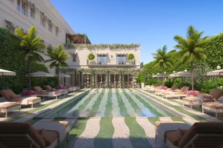 Lujosa zona de piscina al aire libre con azulejos a rayas, rodeada de tumbonas, sombrillas, palmeras y un edificio adornado con vegetación.