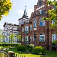 Historische deutsche Villa mit roter Backsteinfassade, markantem Rundturm mit kegelförmigem Dach und Fahne, Fachwerkelementen, gepflegten Gärten mit geschnittenen Hecken, altem Baumbestand und traditionellem Zaun unter blauem Himmel.