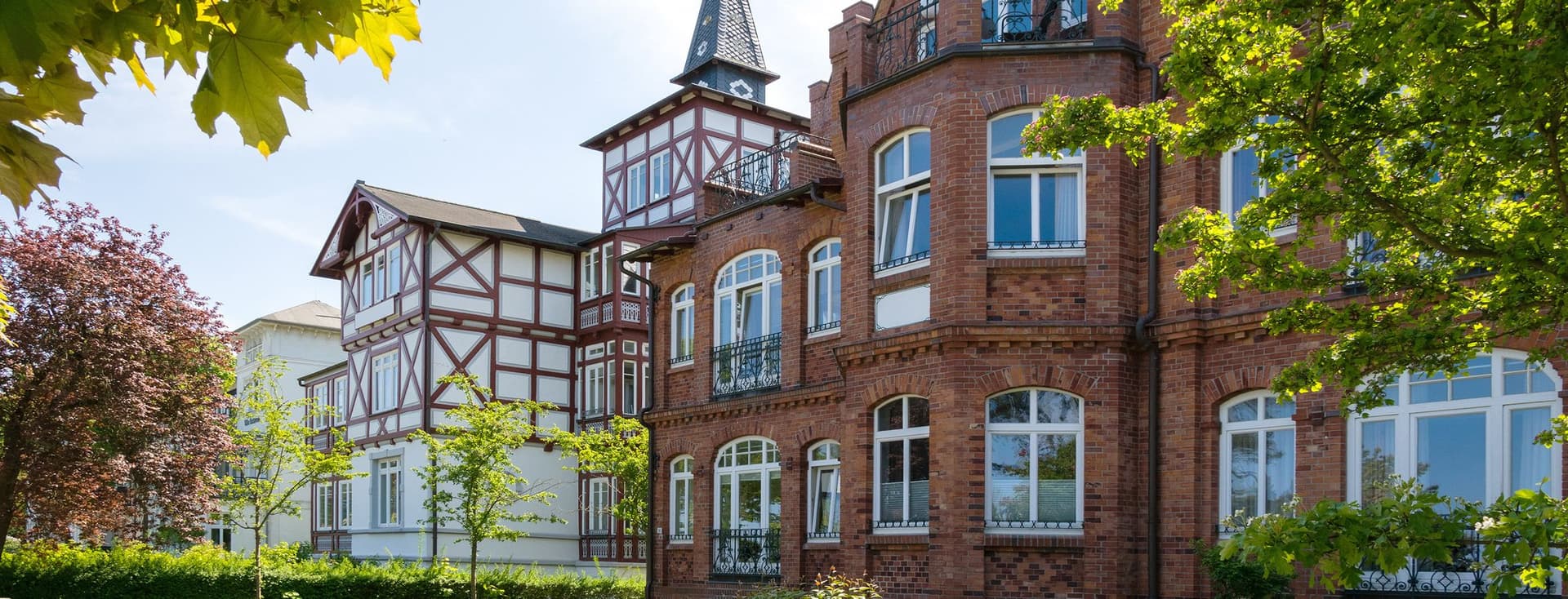 Historic German villa featuring red brick construction, distinctive round tower with conical roof and flag, half-timbered elements, landscaped gardens with manicured hedges, mature trees, and traditional fence under blue sky