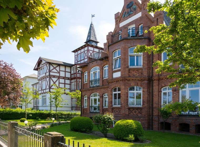 Historic German villa featuring red brick construction, distinctive round tower with conical roof and flag, half-timbered elements, landscaped gardens with manicured hedges, mature trees, and traditional fence under blue sky