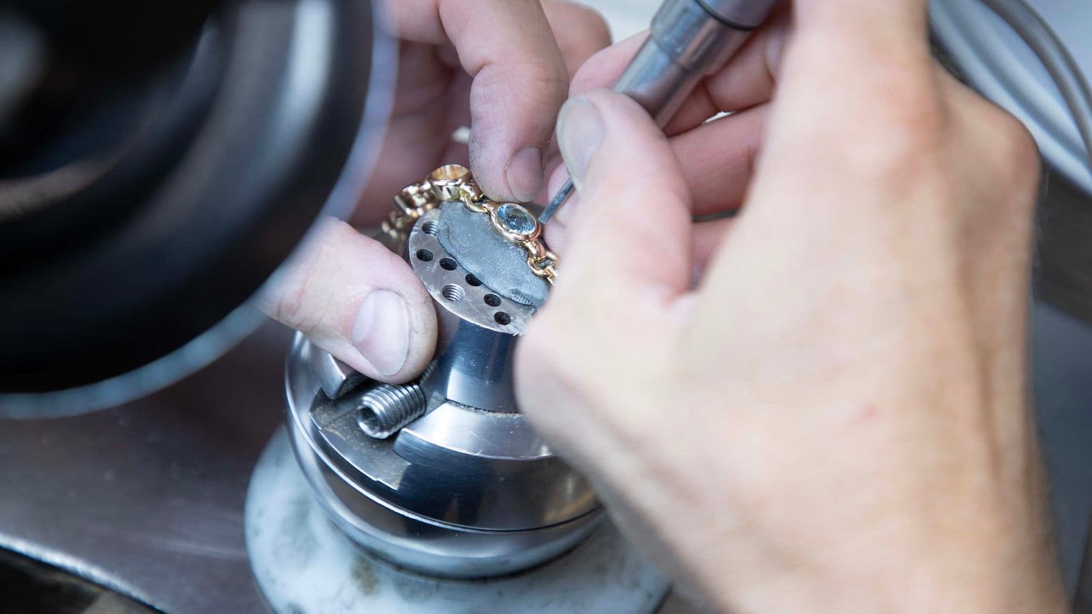 Close-up of jeweler's hands using a tool on a gold bracelet with blue stones, held in a metal clamp.