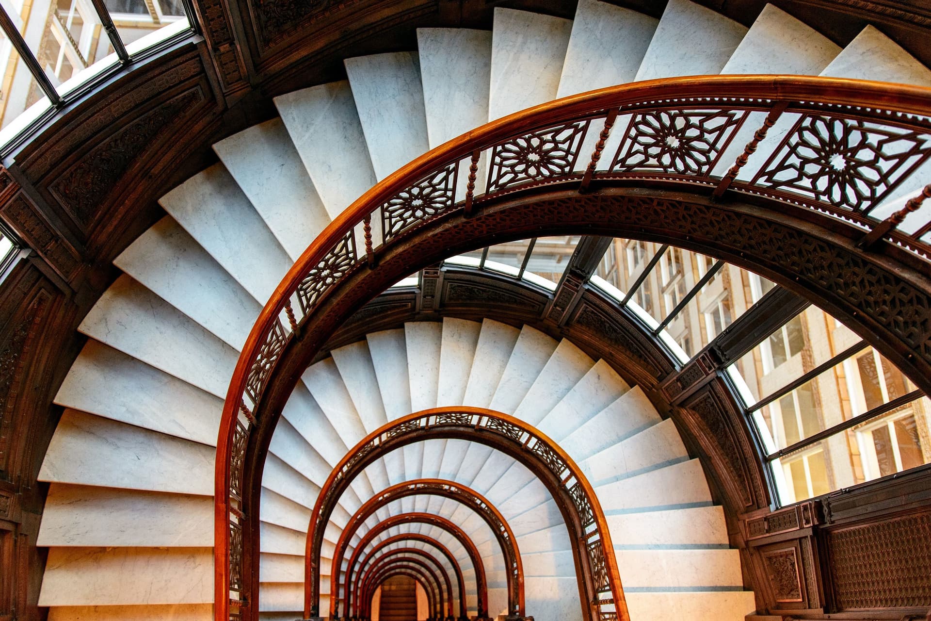 A high-angle shot of a spiral staircase with white marble steps and ornate wood railings.