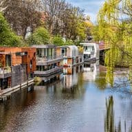 Eine Reihe moderner Hausboote mit zeitgenössischer Architektur, darunter Holzverkleidungen und große Fenster, die entlang eines von Bäumen gesäumten Kanals mit Trauerweiden, ruhigen Wasserreflexionen und üppigem grünem Frühlingslaub vor Anker liegen.