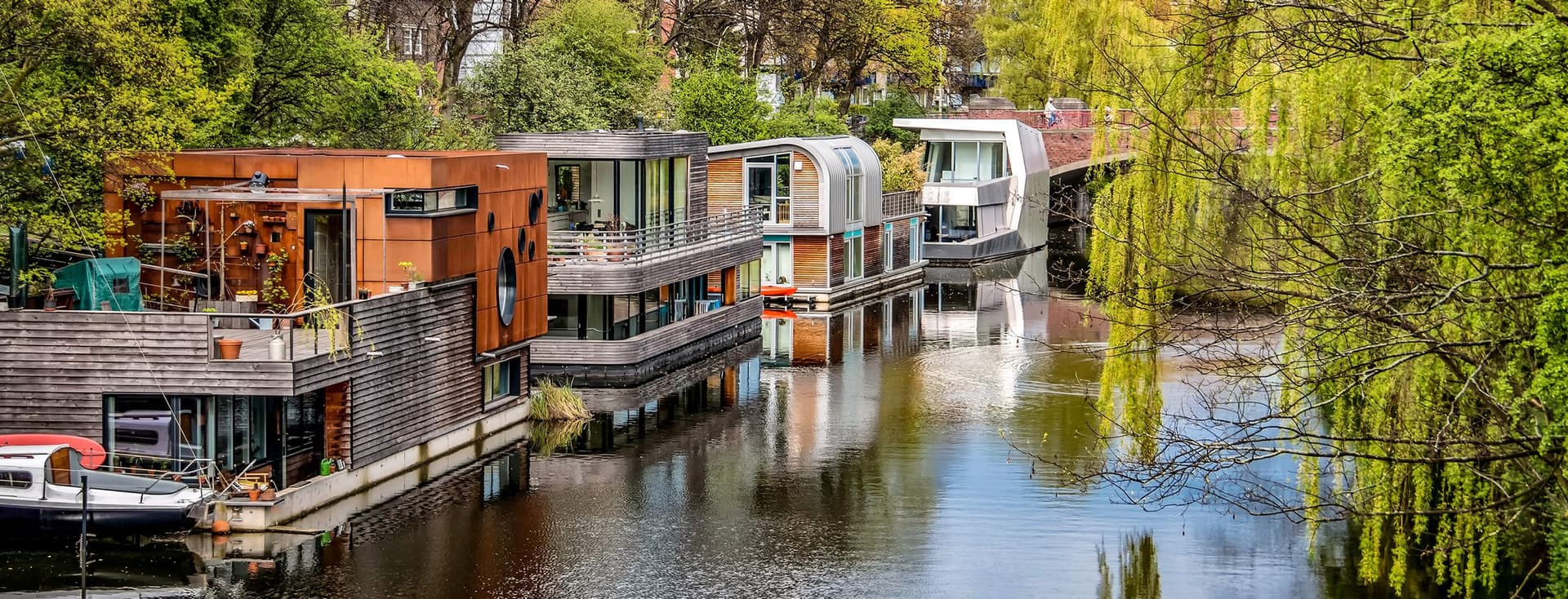 Row of modern houseboats with contemporary architecture including wood cladding and large windows moored along tree-lined canal with weeping willows, calm water reflections, and lush green spring foliage