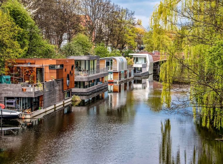 Row of modern houseboats with contemporary architecture including wood cladding and large windows moored along tree-lined canal with weeping willows, calm water reflections, and lush green spring foliage