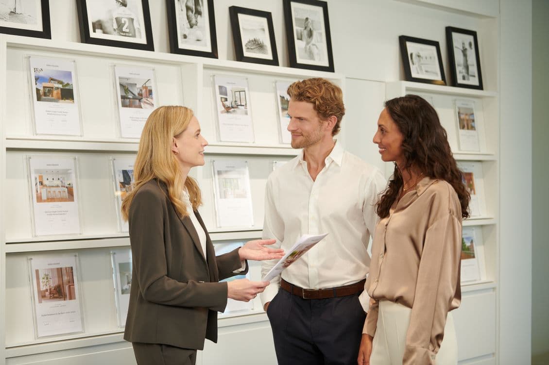 A real estate agent shows a brochure to a smiling couple in an office with property listings displayed on the wall.
