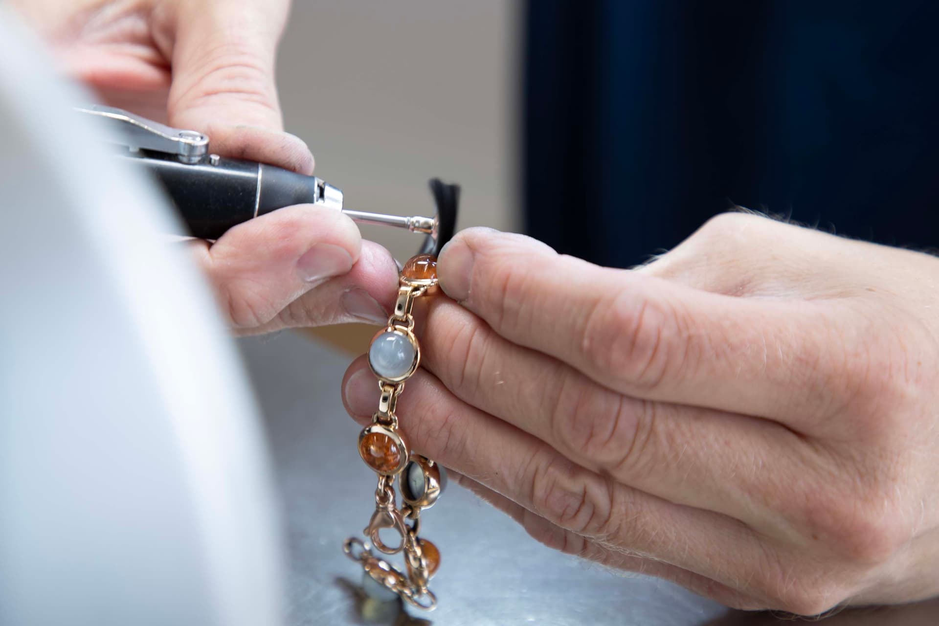 Close-up of hands polishing a gold bracelet with amber and moonstone beads using a small black power tool.