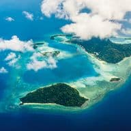 Luftaufnahme einer tropischen Insel mit üppiger grüner Vegetation, umgeben von türkisfarbenem Wasser und weißen Wolken.