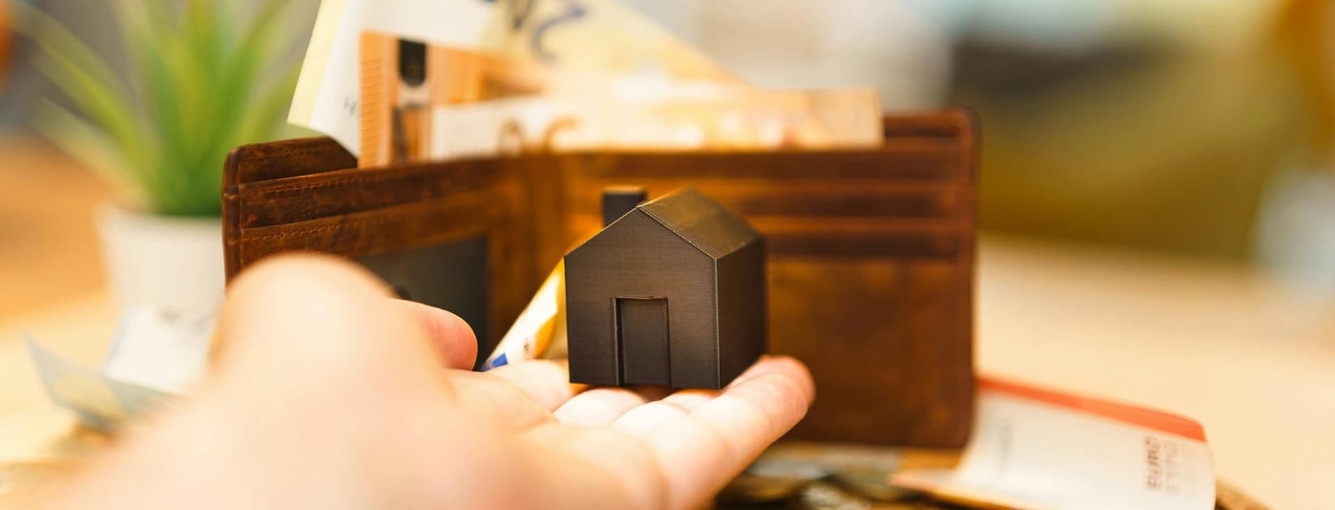Ένα χέρι κρατά ένα μικρό ειδώλιο σπιτιού μπροστά από ένα ανοιχτό πορτοφόλι γεμάτο με τραπεζογραμμάτια ευρώ, κέρματα και πιστωτικές κάρτες, που συμβολίζουν τα οικονομικά και τα ακίνητα.