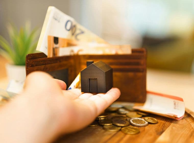 Ένα χέρι κρατά ένα μικρό ειδώλιο σπιτιού μπροστά από ένα ανοιχτό πορτοφόλι γεμάτο με τραπεζογραμμάτια ευρώ, κέρματα και πιστωτικές κάρτες, που συμβολίζουν τα οικονομικά και τα ακίνητα.