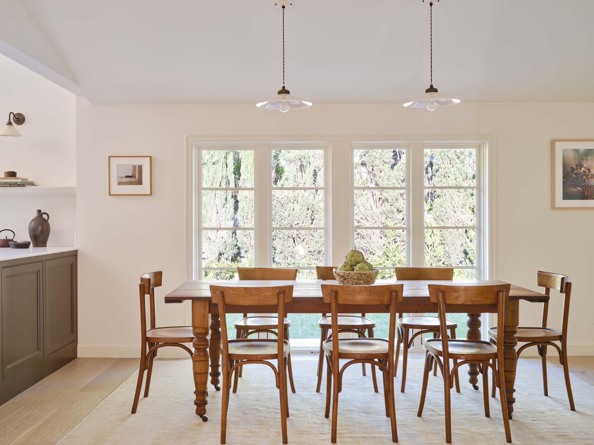 Bright dining room with a wooden table, chairs, and a basket of artichokes. Large windows overlook green trees.