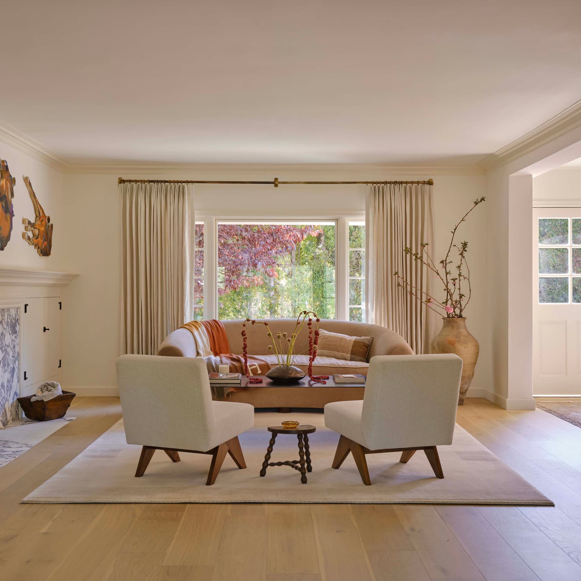 Living room with a beige sofa, two chairs, and a coffee table on a light rug. A fireplace is on the left and a window with curtains is in the background.