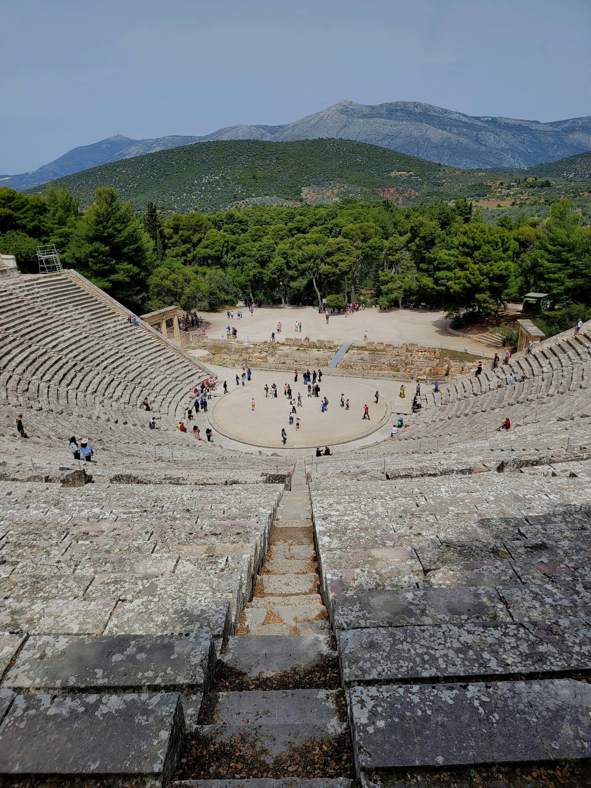 View of the Ancient Theatre of Epidaurus, Greece. Stone seating surrounds a circular stage, with green trees and mountains in the background. People are scattered throughout the theater.