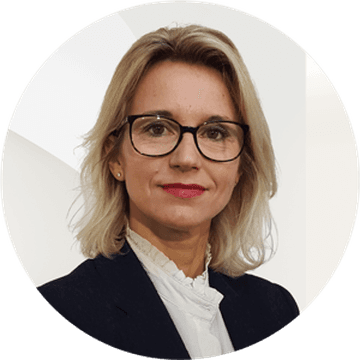 Portrait d'une femme blonde portant des lunettes, vêtue d'un blazer foncé et d'un chemisier blanc, sur fond blanc.