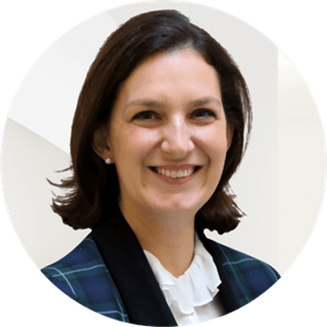 Une femme souriante aux cheveux courts bruns porte un blazer à carreaux bleus et verts sur un chemisier blanc à volants.